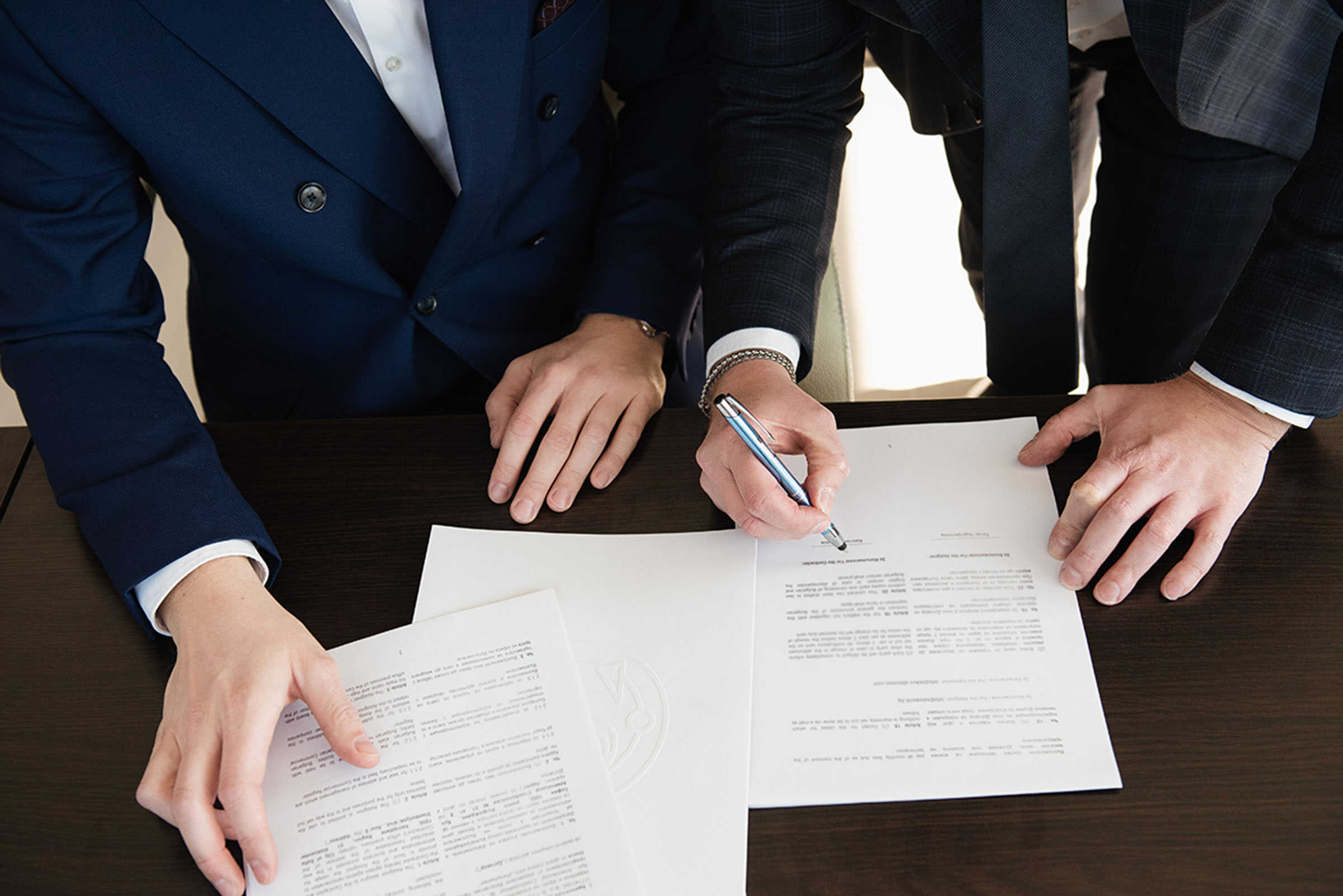 Alt text: Two professionals in suits signing legal documents at a meeting.